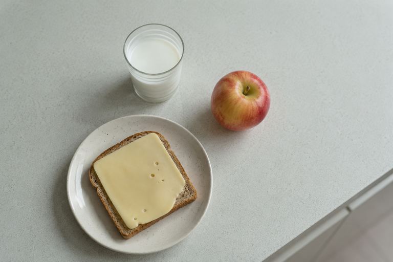 Een boterham met kaas op een bordje, met een glas melk en een appel op een keukenblad.